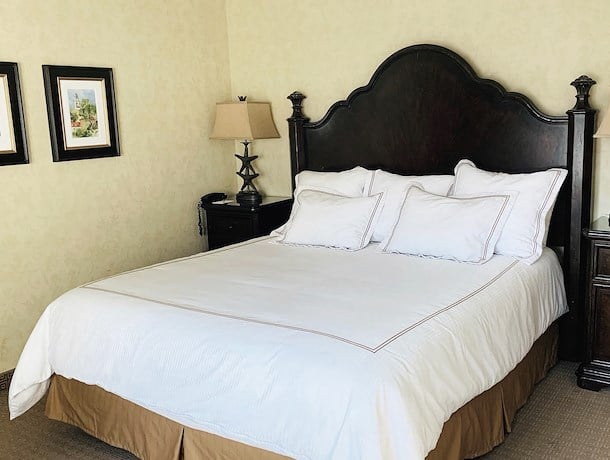 A neatly made bed with white linens and a dark wooden headboard in a room with beige walls.