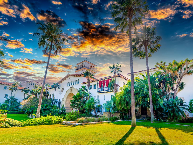 A large white building with Spanish-style architecture and palm trees in front of it, set against a backdrop of a colorful sunset.